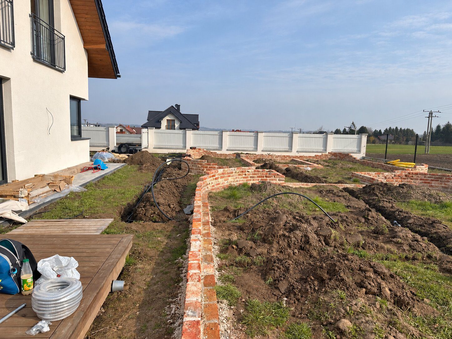 Ogród w trakcie budowy: wymurowane ceglane obrzeża rabat, rozkopana ziemia, rozłożone rury nawadniania i drewniany taras przy domu